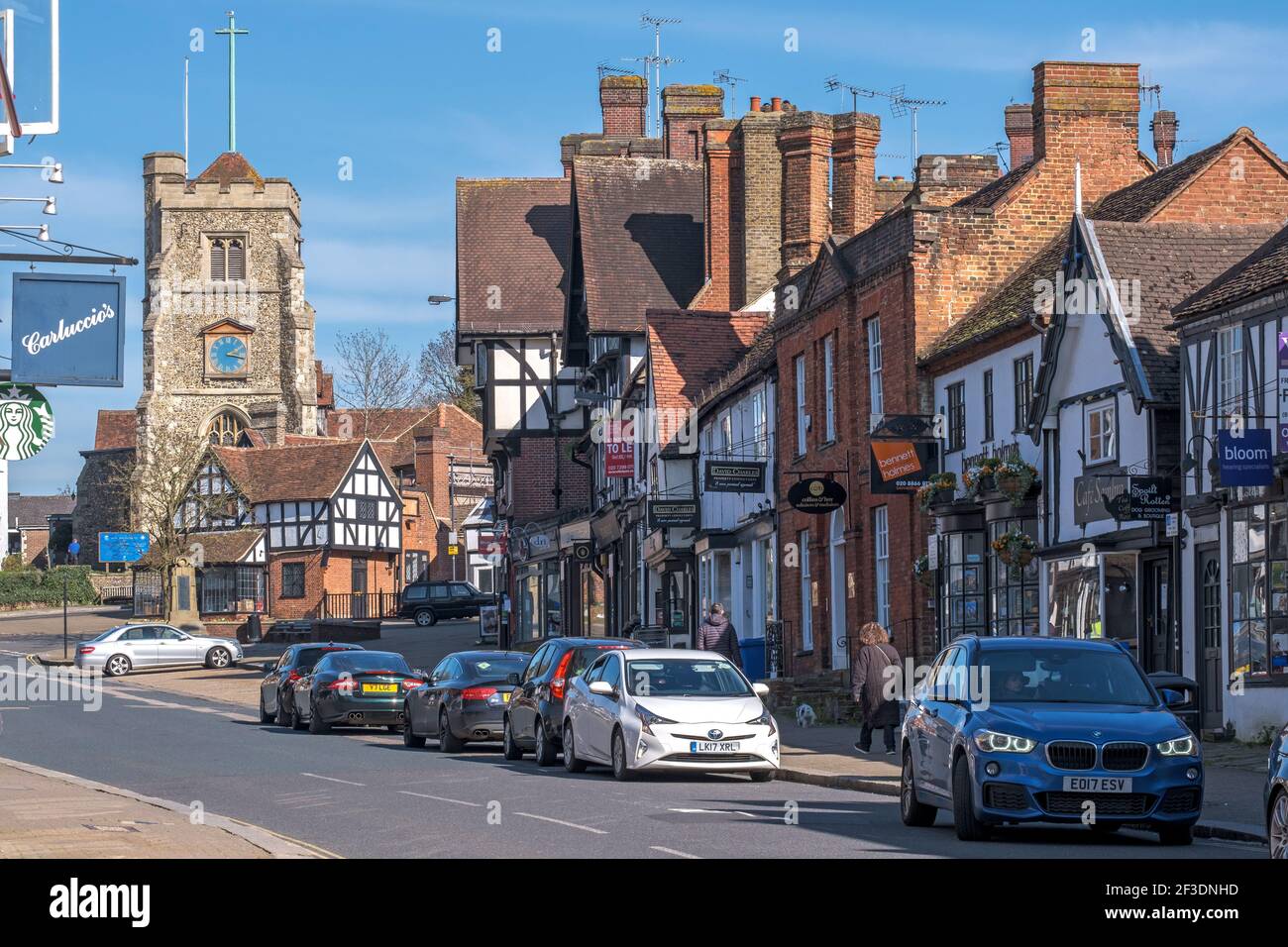 Pinner Village high street with the medieval hilltop church St John the Baptist, shops, parked cars and two people walking on the pavement. England Stock Photo