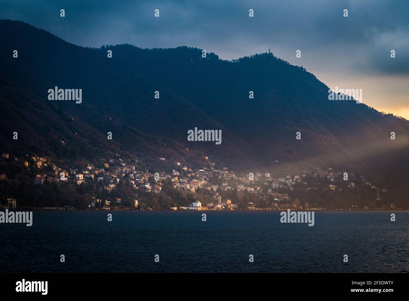 Cropped photo of village in wooded slope on opposite shore of lake. Buildings illuminated by rays of sun. Stock Photo