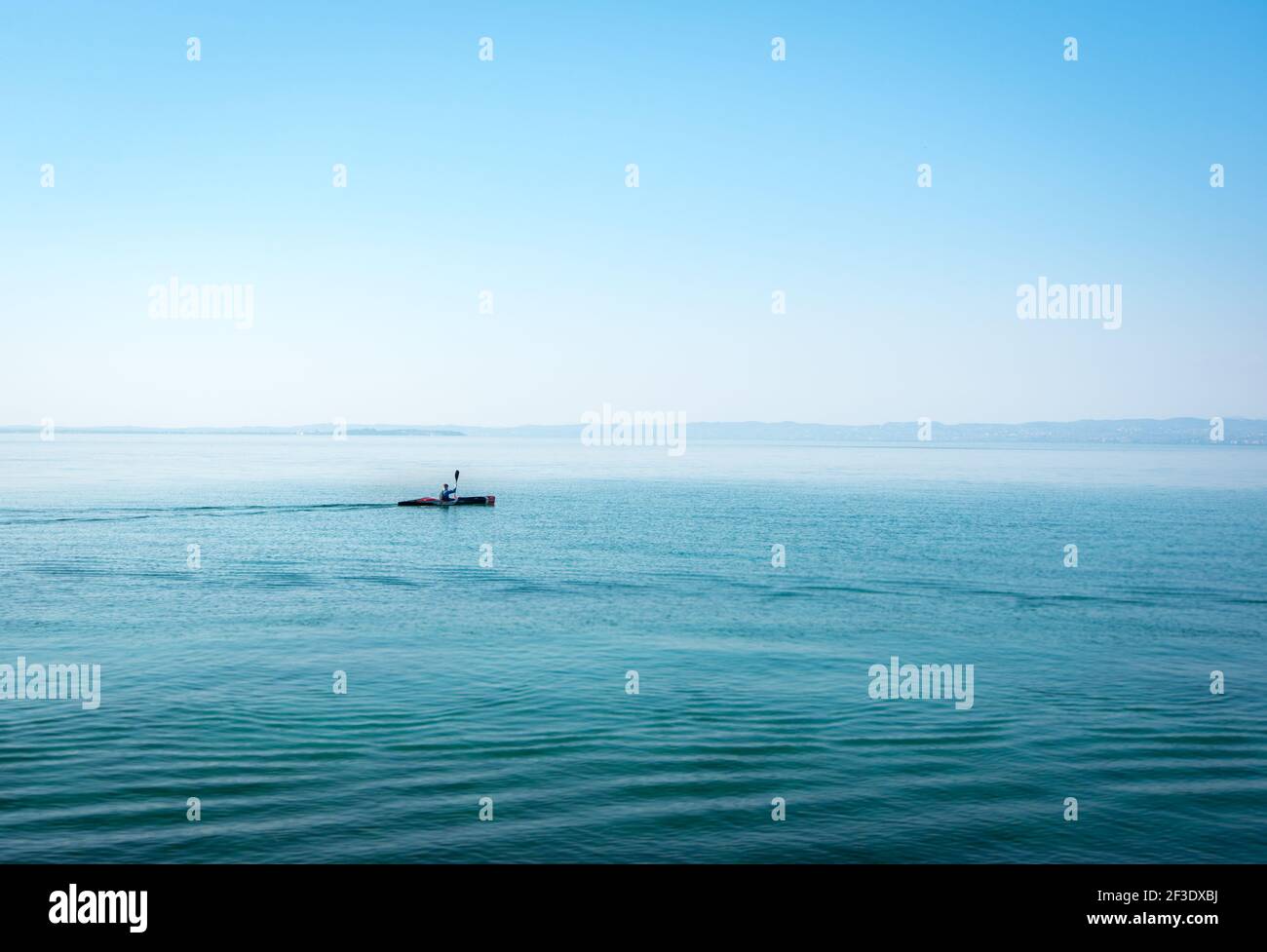 Alone sportsman in kayak on wide water surface - Garda Lake. Single person paddling on calm lake in morning. Clear sky. Stock Photo