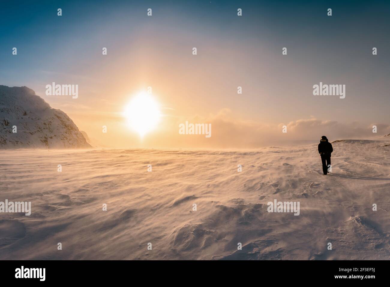 Scenic picture of large Sunset halo in winter mountains, strong snow drift wind. A person in warm black snowmobile overall and goggles walks against s Stock Photo