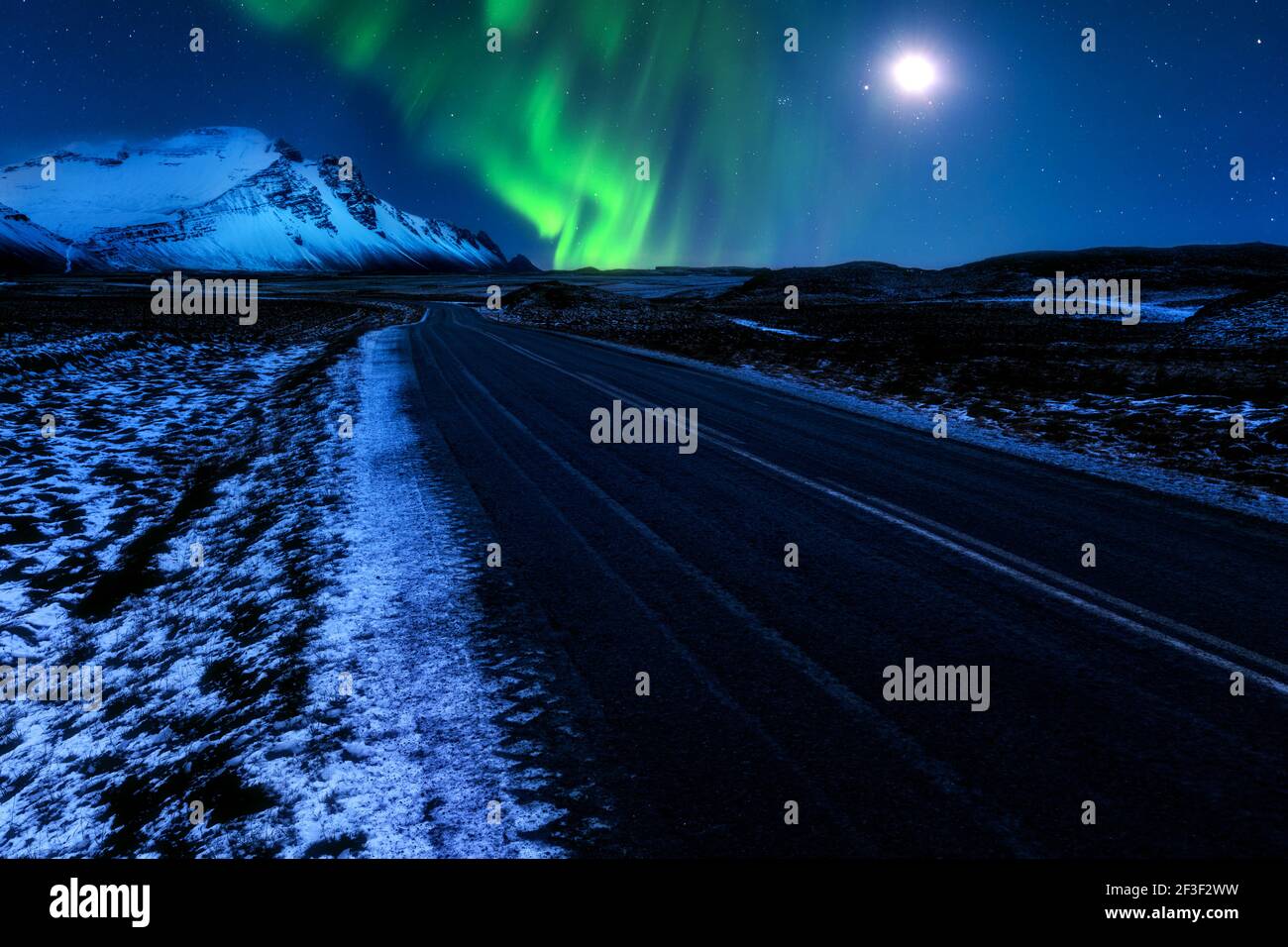 Icelandic Hringvegur ring-road (route 1) leads towards the snow-covered Vestrahorn mountains, below the Northern Lights and the moon. Stock Photo