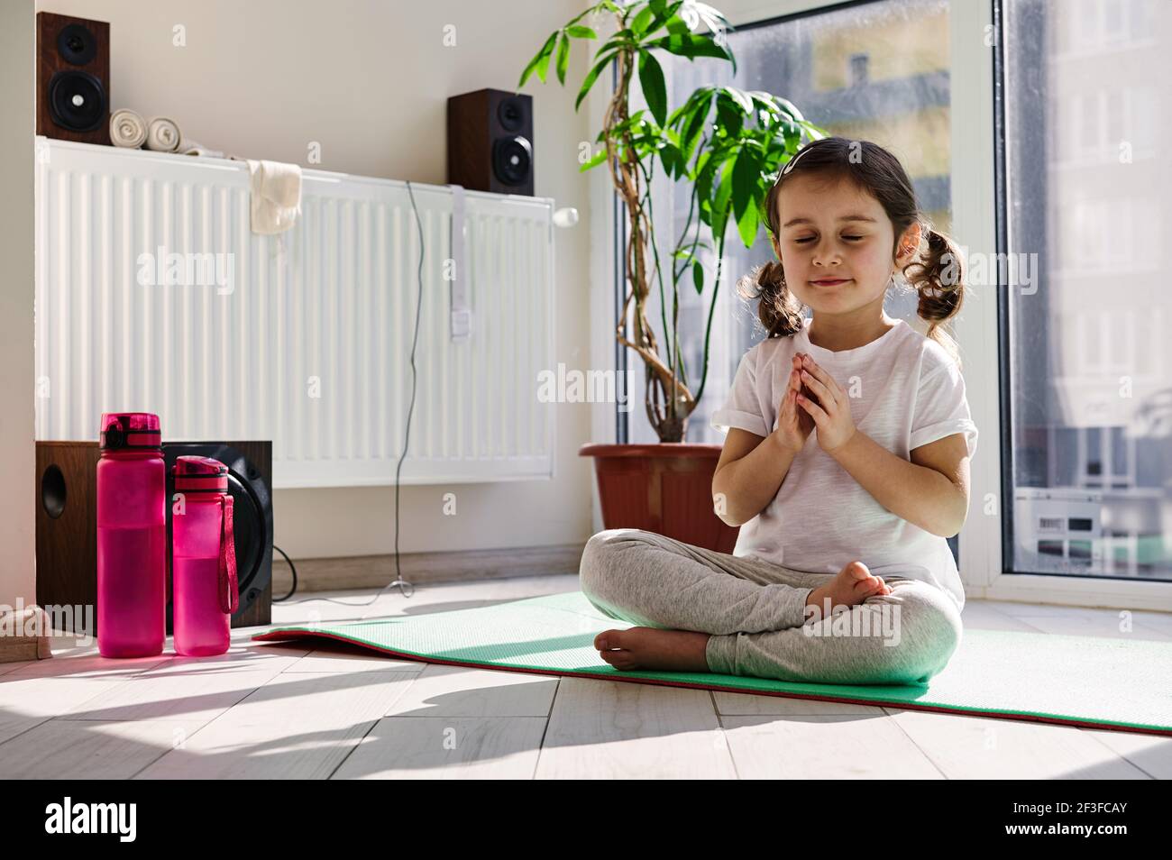 Beautiful little girl sitting on a fitness mat and practicing yoga in lotus position against the background of large windows at home. Proper healthy p Stock Photo