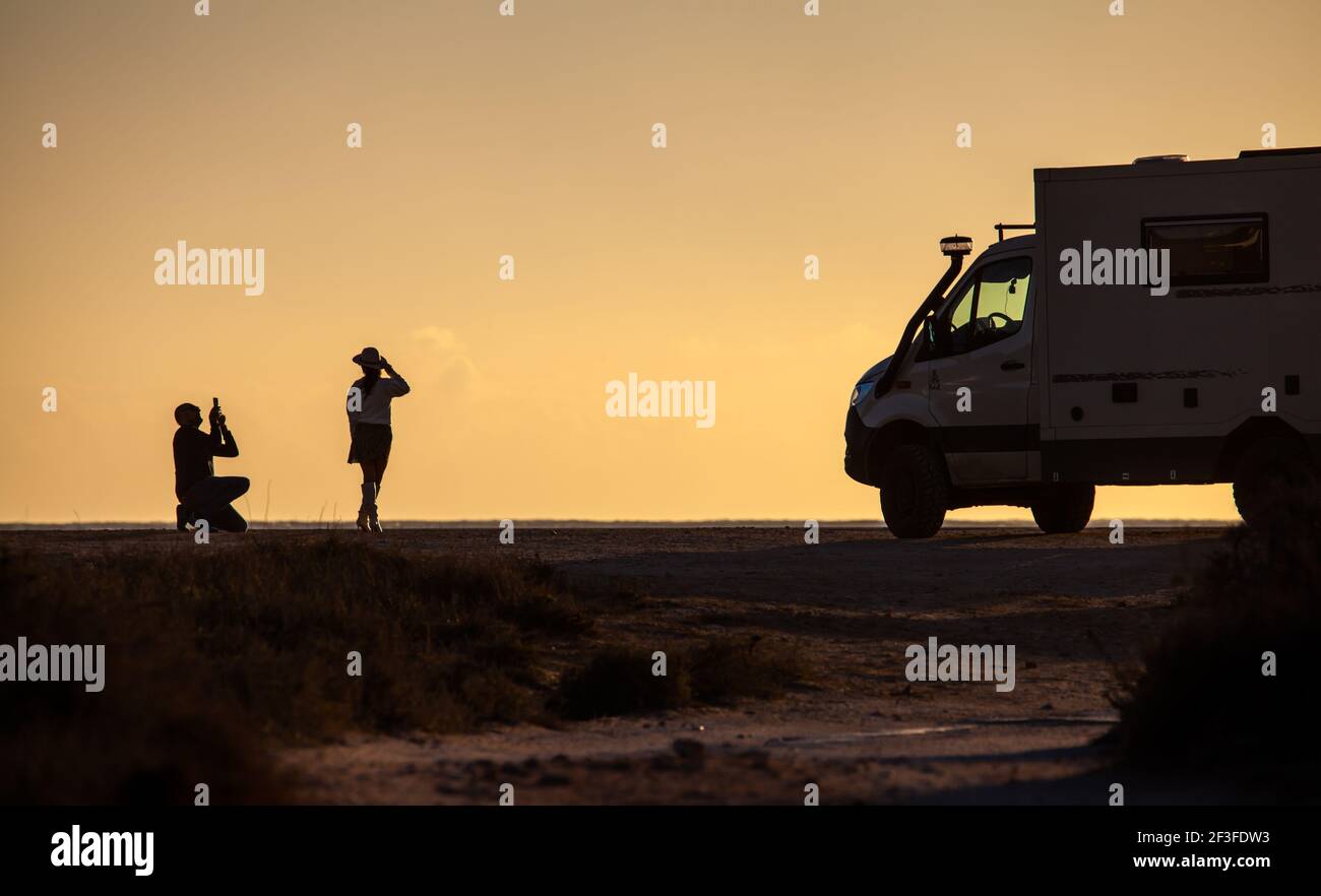 Silhouette of couple taking photos with cellphone in front of offroad motorhome truck in the evening sunlight Stock Photo