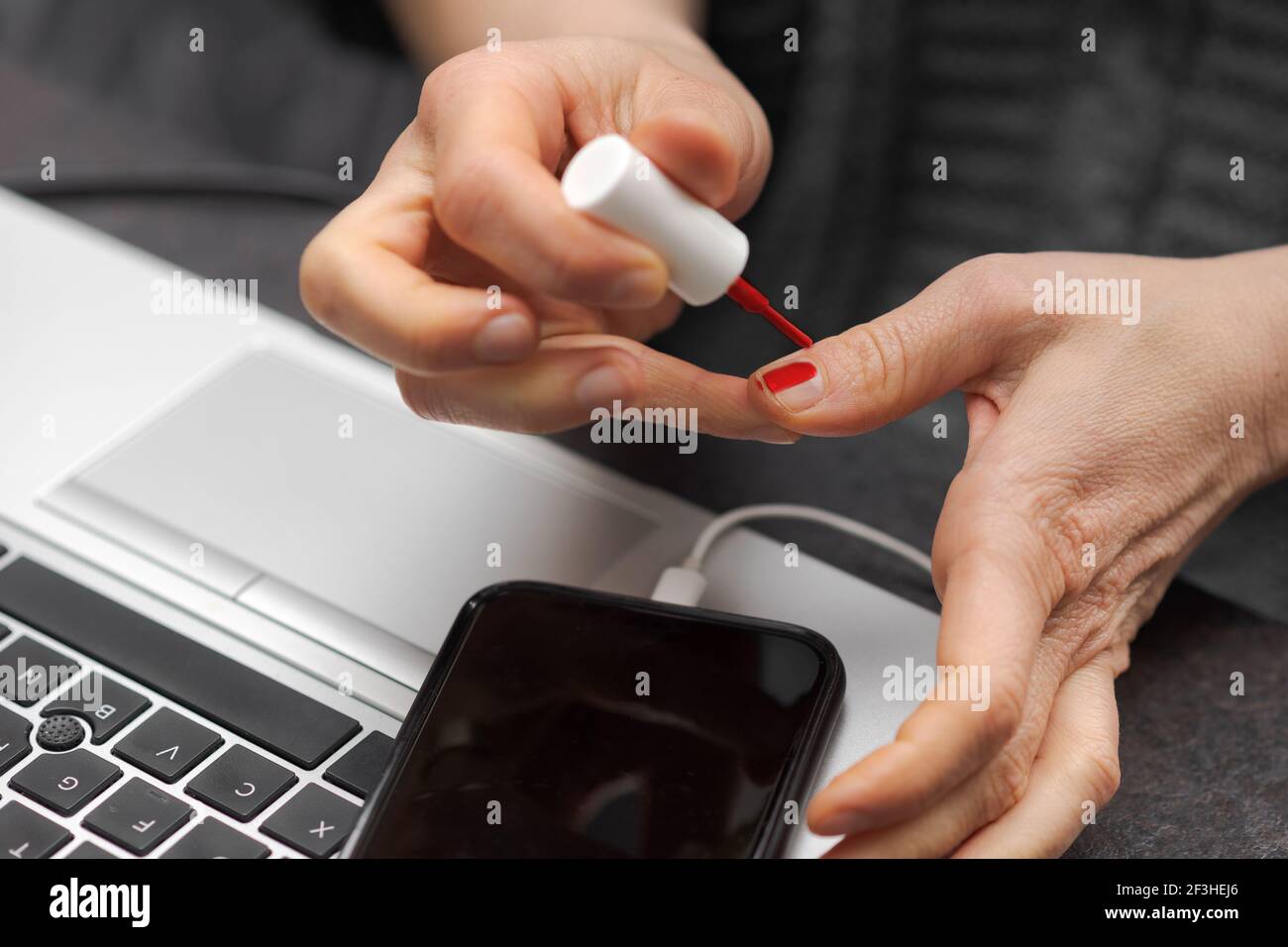 Female or male hands applying red nail polish on fingernails as stress relieve from work at desk in home office near laptop computer and smartphone Stock Photo