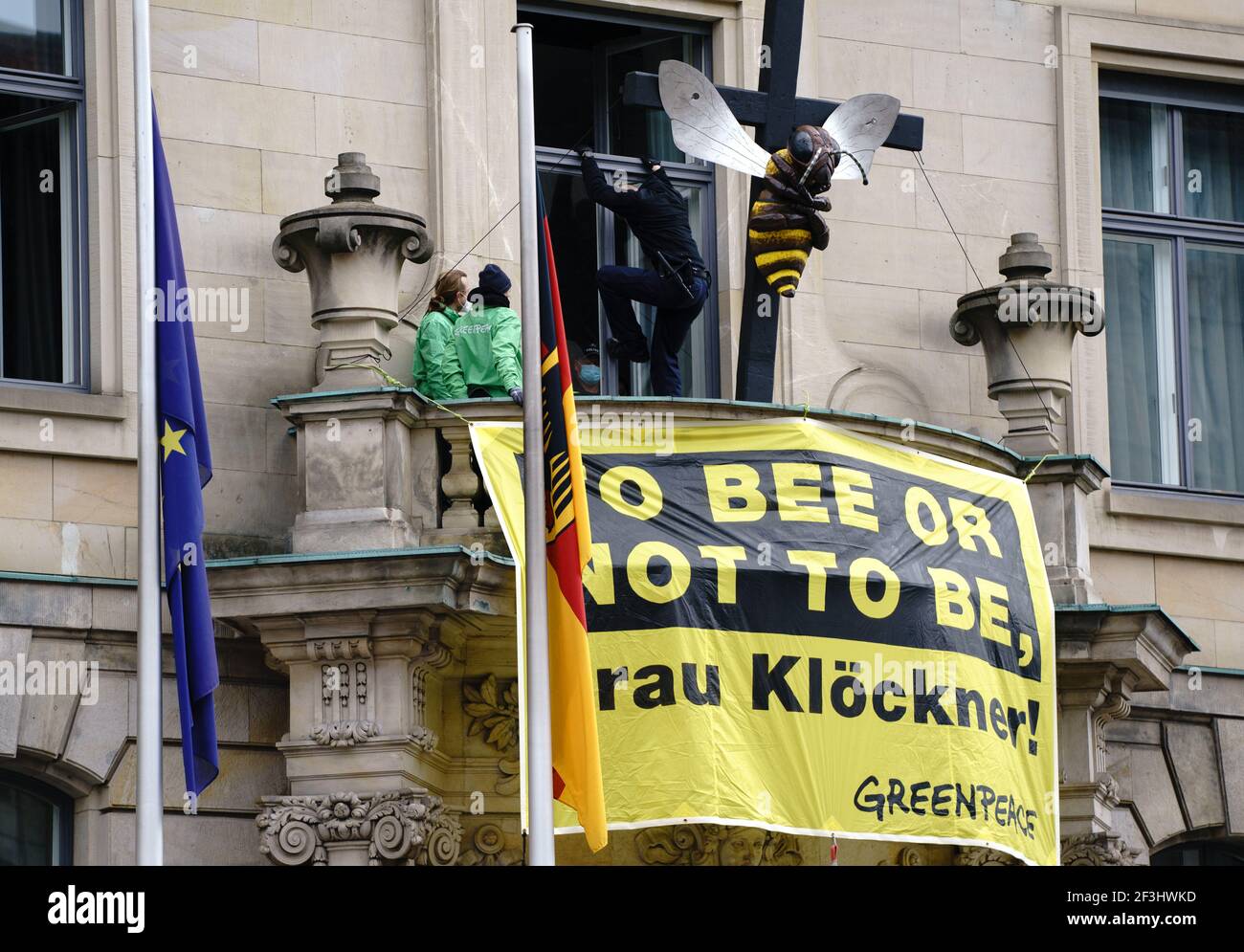 Berlin, Germany. 17th Mar, 2021. Greenpeace activists display a poster with the play on words 'To bee or not to be, Frau Klöckner' in front of their sculpture of a bee on a cross on the balcony of the Federal Ministry of Agriculture, while a police officer climbs through an upper window. With the action, the environmentalists protest for the preservation of diversity in the fields and demand more funding for species protection. Credit: Kay Nietfeld/dpa/Alamy Live News Stock Photo