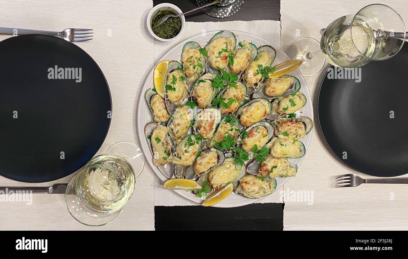 halves of New Zealand green Perna canaliculus mussels baked with cream sauce and cheese on a platter, decorated with lemon wedges. Two glasses of whit Stock Photo