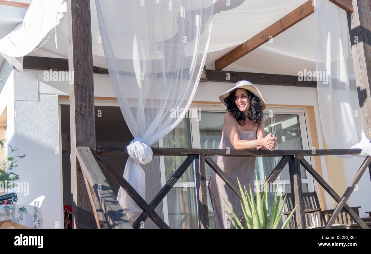 smiling pretty young brunette woman with curly hair and hat contemplates the morning from the balcony of her vacation chalet in summer illuminated by Stock Photo