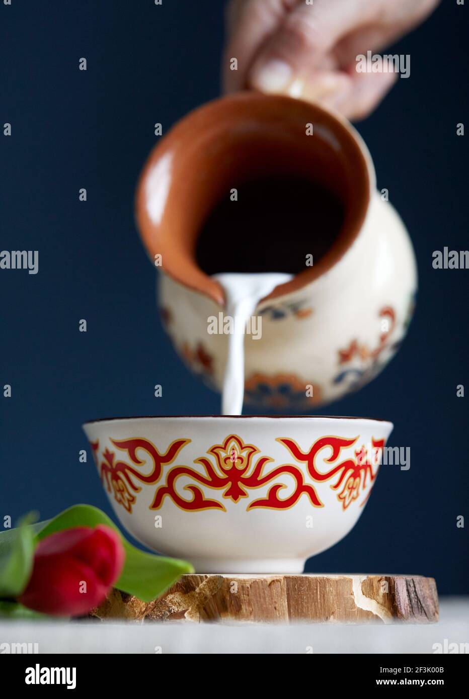 Kazakh woman pouring out milk in traditional tea bowl kese with Kazakh ornament near read tulips at dark blue background during Nauryz festival. Stock Photo