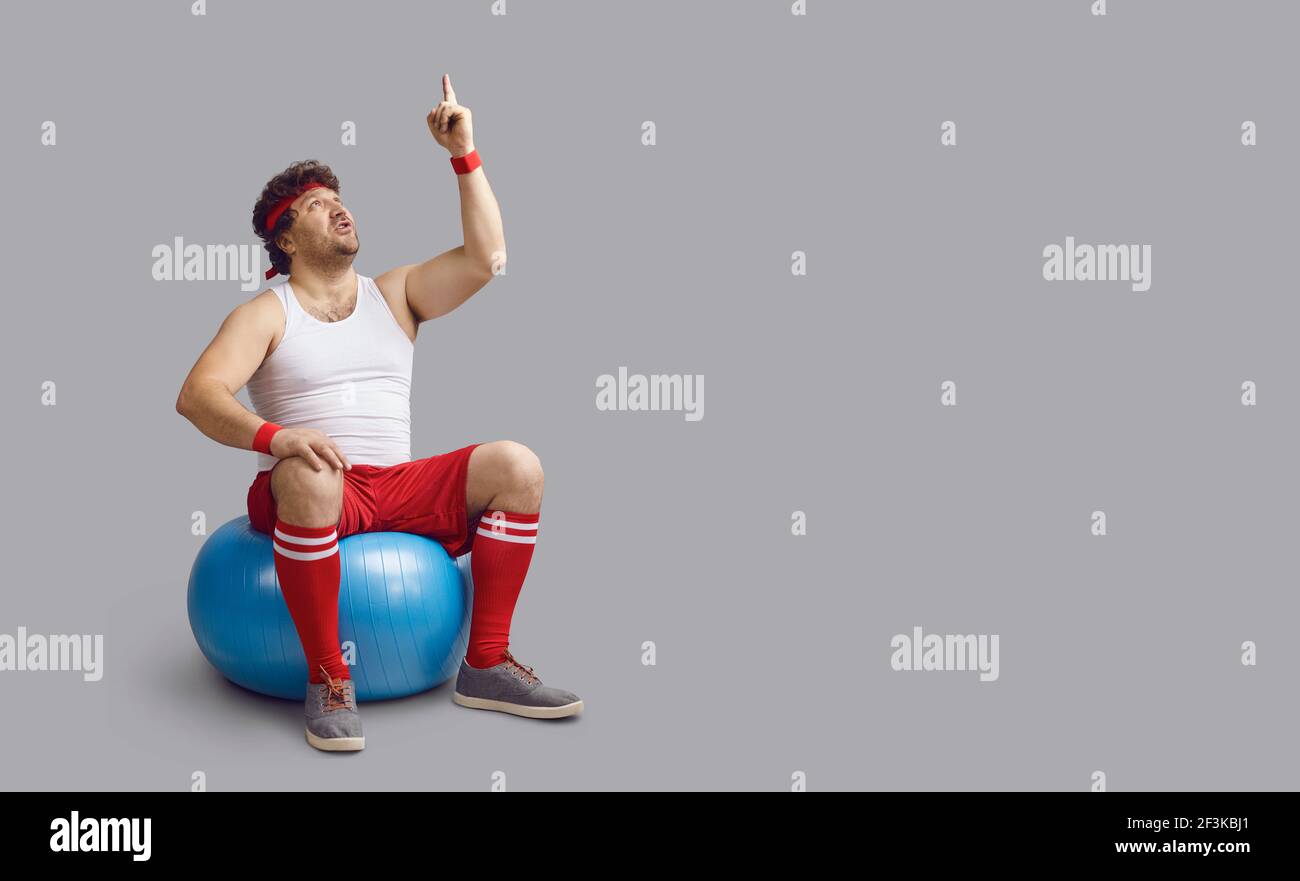 Funny chubby man sitting on fit ball, looking up and pointing at something above Stock Photo