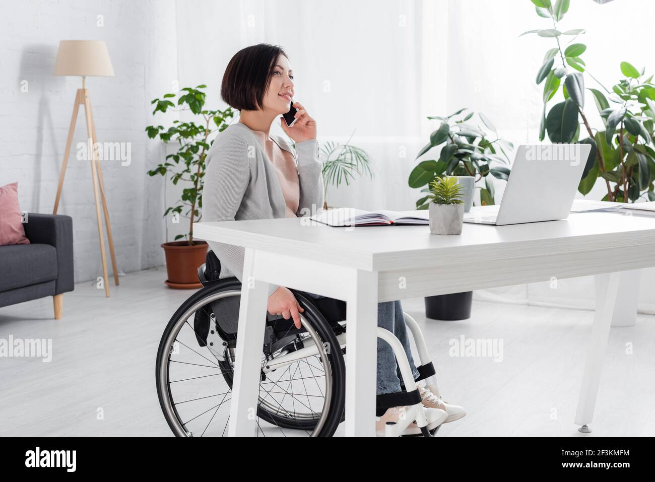 Smiling handicapped woman talking on smartphone near laptop and notebook at home Stock Photo