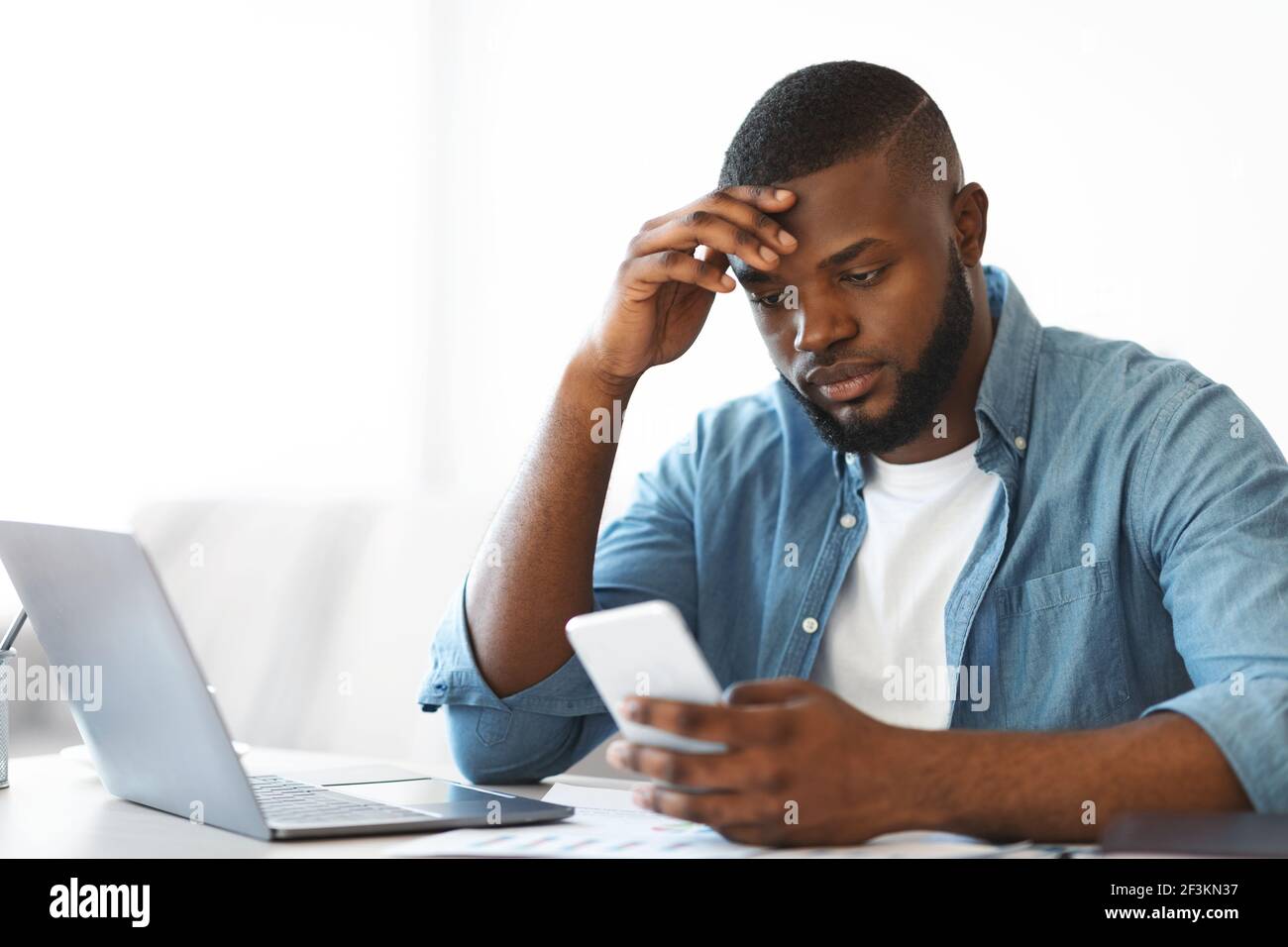 Concerned black freelancer guy looking at smartphone while working at home office Stock Photo