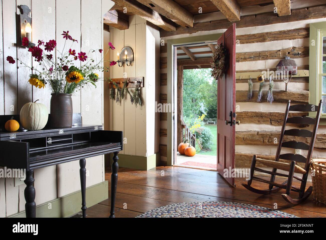 Front door and hall area of historic log cabin, with exposed logs and rocking chair. Also exposed beams, hardwood floors and fall decor and flowers. Stock Photo
