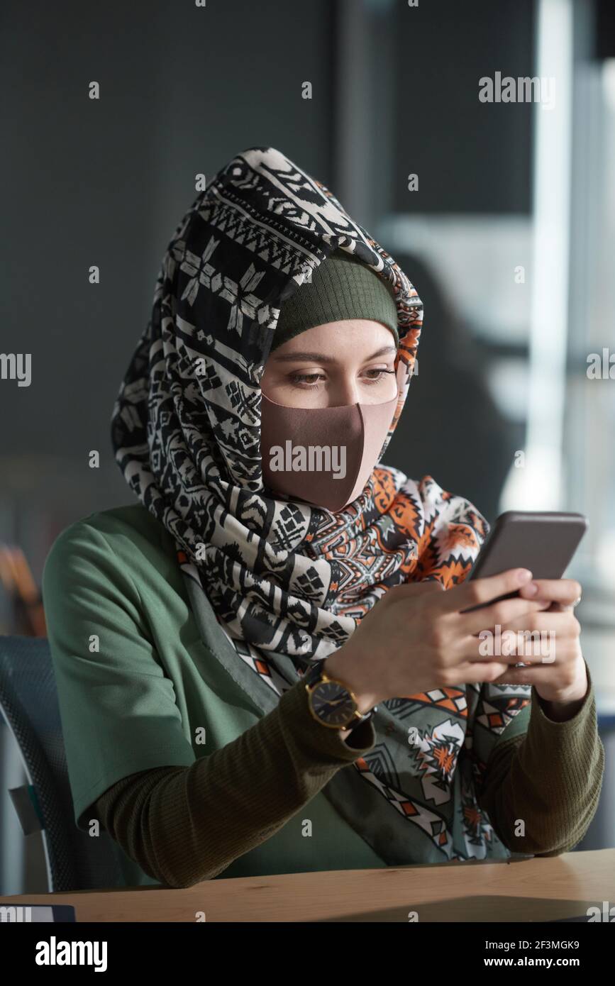 Young muslim woman in hijab and protective mask sitting at the table and using mobile phone Stock Photo