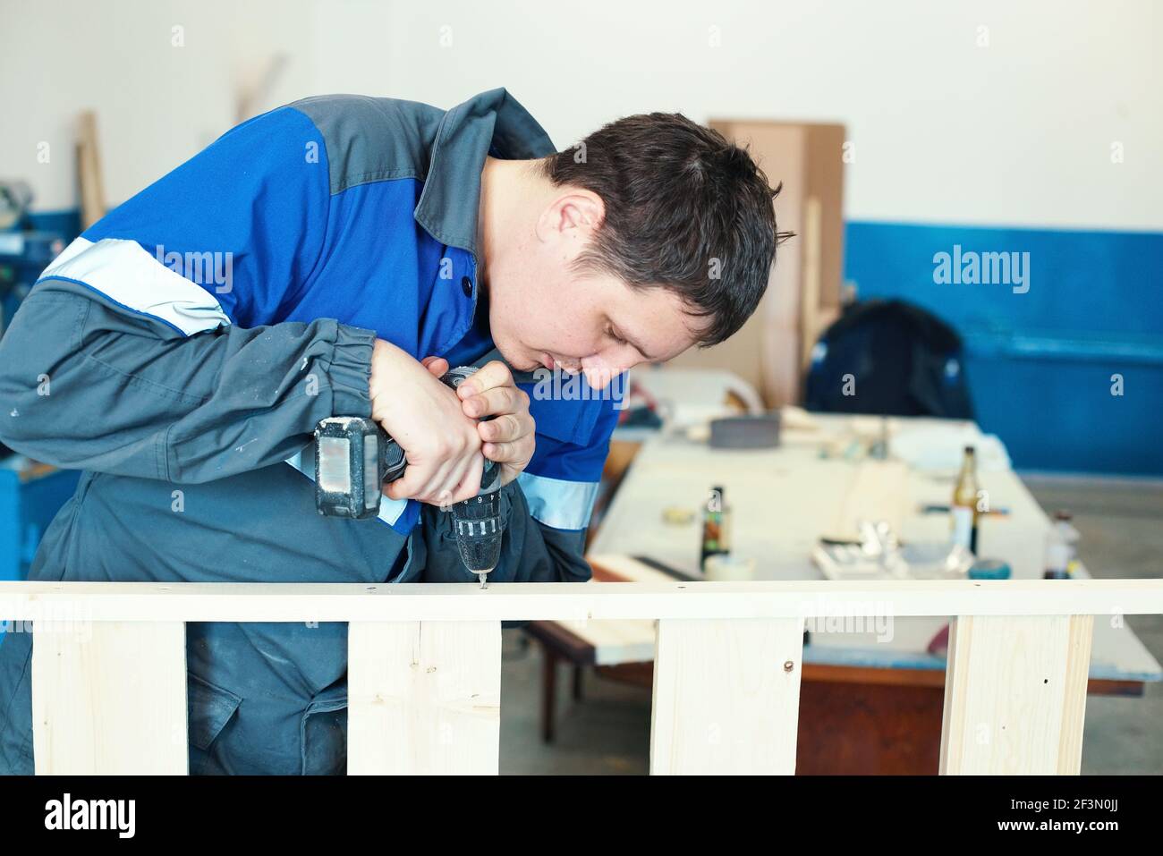 A young worker in overalls with a screwdriver collects furniture in a carpenter's shop. Background for making furniture. Stock Photo