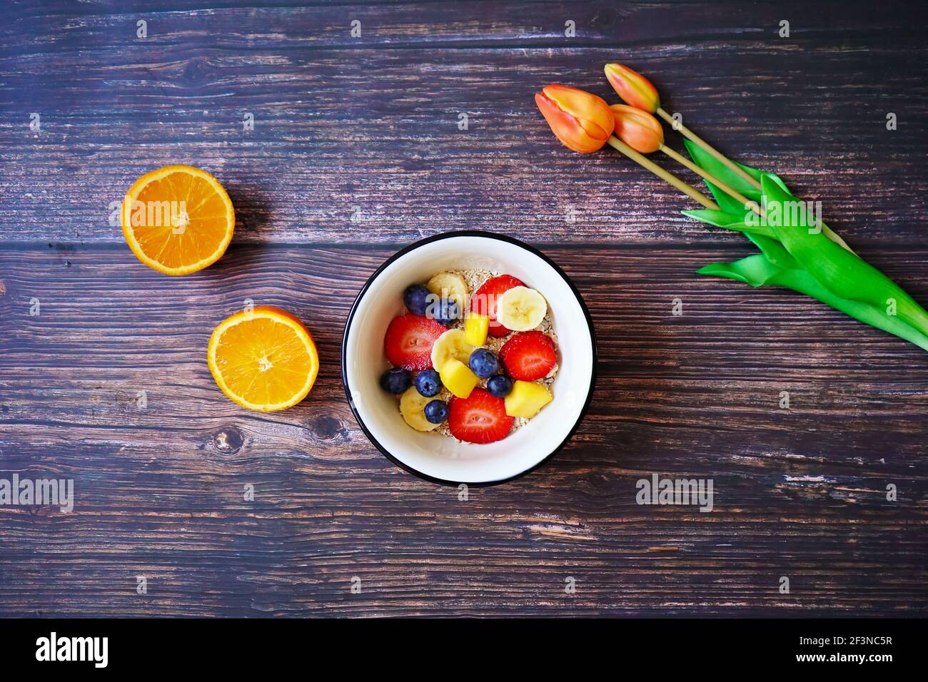 A cereal bowl with fresh fruits (strawberry, banana, blueberry, mango) and an orange on a wooden breakfast table. Orange tulips for decoration. Stock Photo