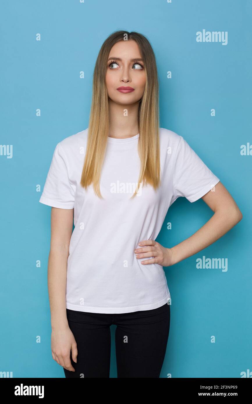 Serious young woman in whte shirt is holding hand on hip and looking aside. Three quarter length studio shot on blue background. Stock Photo