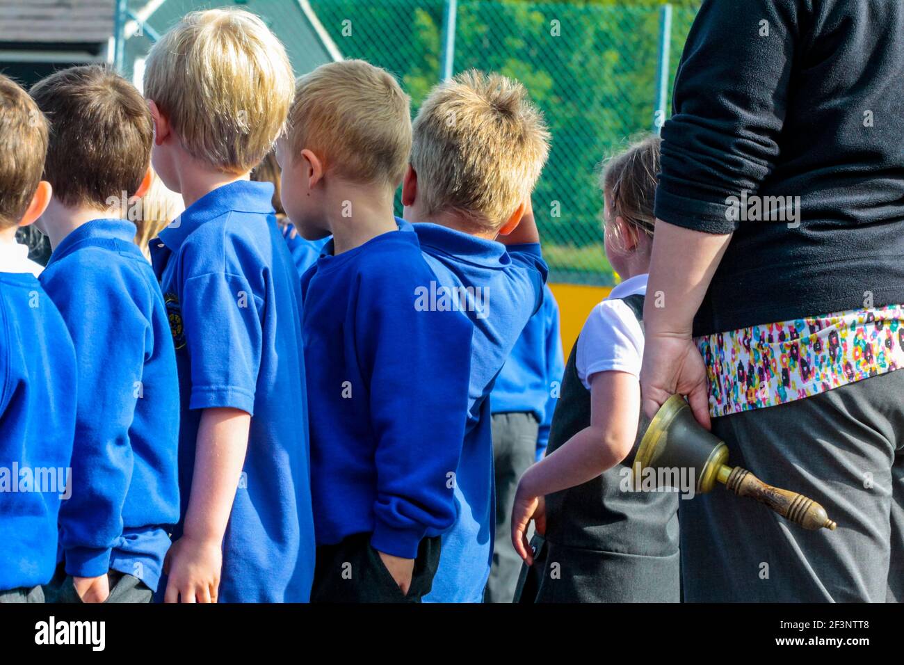 Group of primary school children in school uniform making their way back to the classroom after a break in the lessons next to a teacher holding bell. Stock Photo