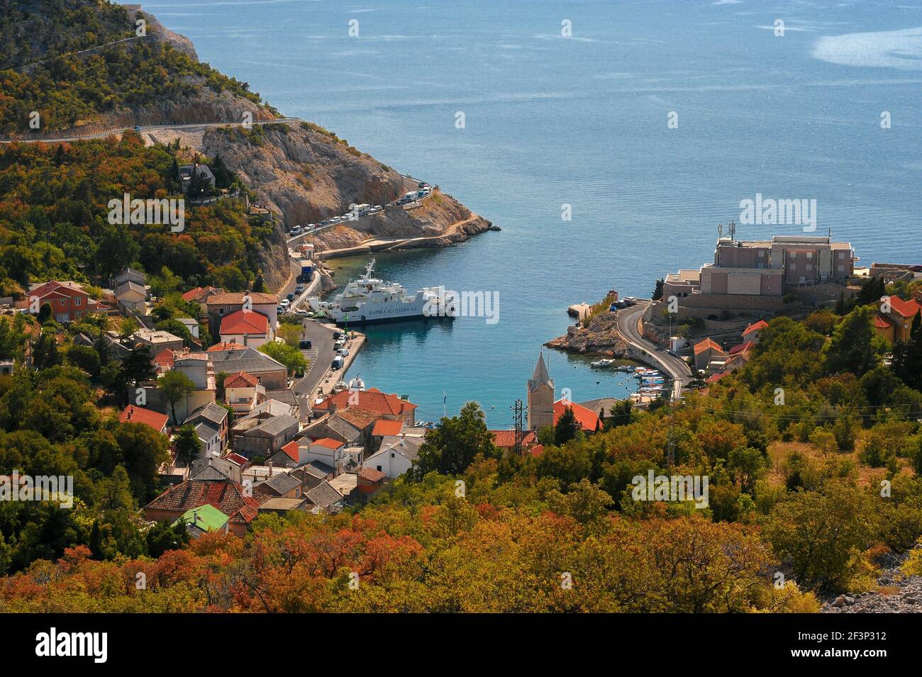 Lower slopes of the 1,757 m / 5,764 ft Velebit mountain range glow with vibrant early autumn oranges and yellow-greens as tourist cars, caravans and motorhomes queue on the narrow corniche road winding down to the tiny Adriatic harbour at Jablanac, Lika-Senj County, western Croatia, to board a ferry for the short voyage to the offshore island of Rab.  This image was captured prior to 2012, when the Rapska Plovidba ferry service moved from Jablanac to the ferry port at Stinica, a short distance up the mainland coast. Stock Photo