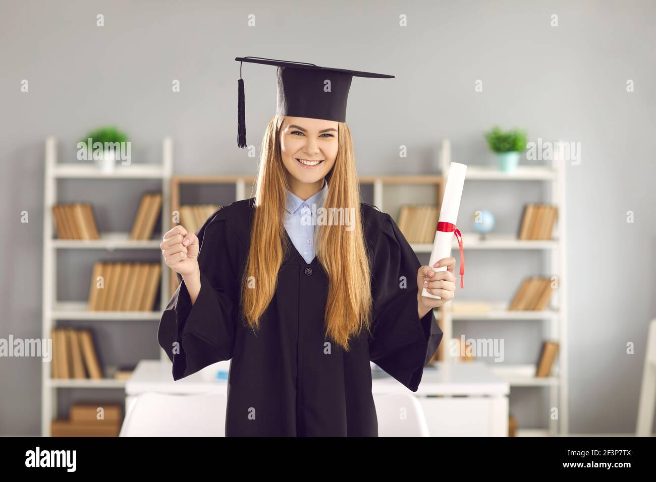 Female graduate holding a diploma in one hand and clenching her fist in joy with the other. Stock Photo