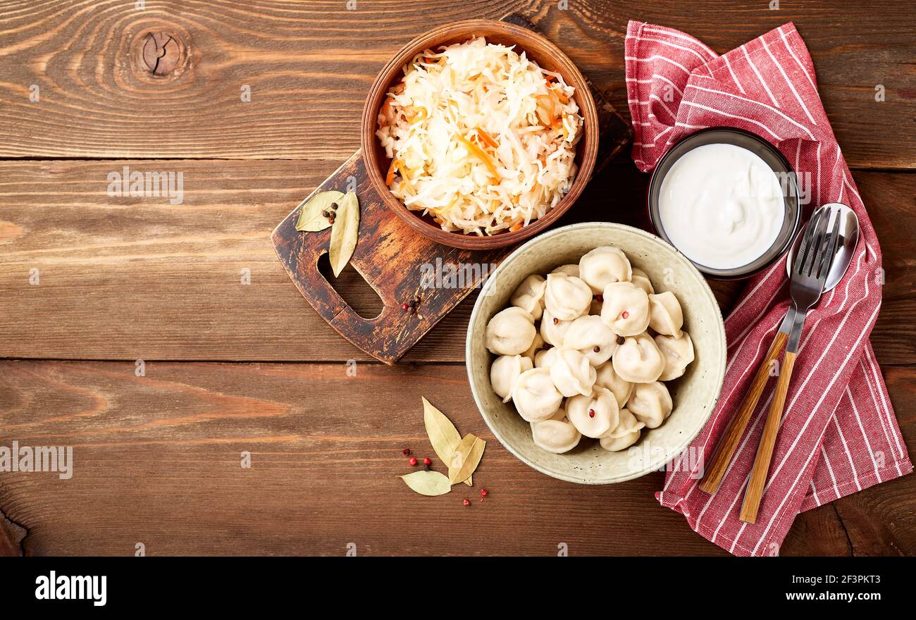 Traditional Russian pelmeni dumplings with sour cream and sauerkraut with sour cream over wooden background. Top view, flat lay. Place for text. Stock Photo