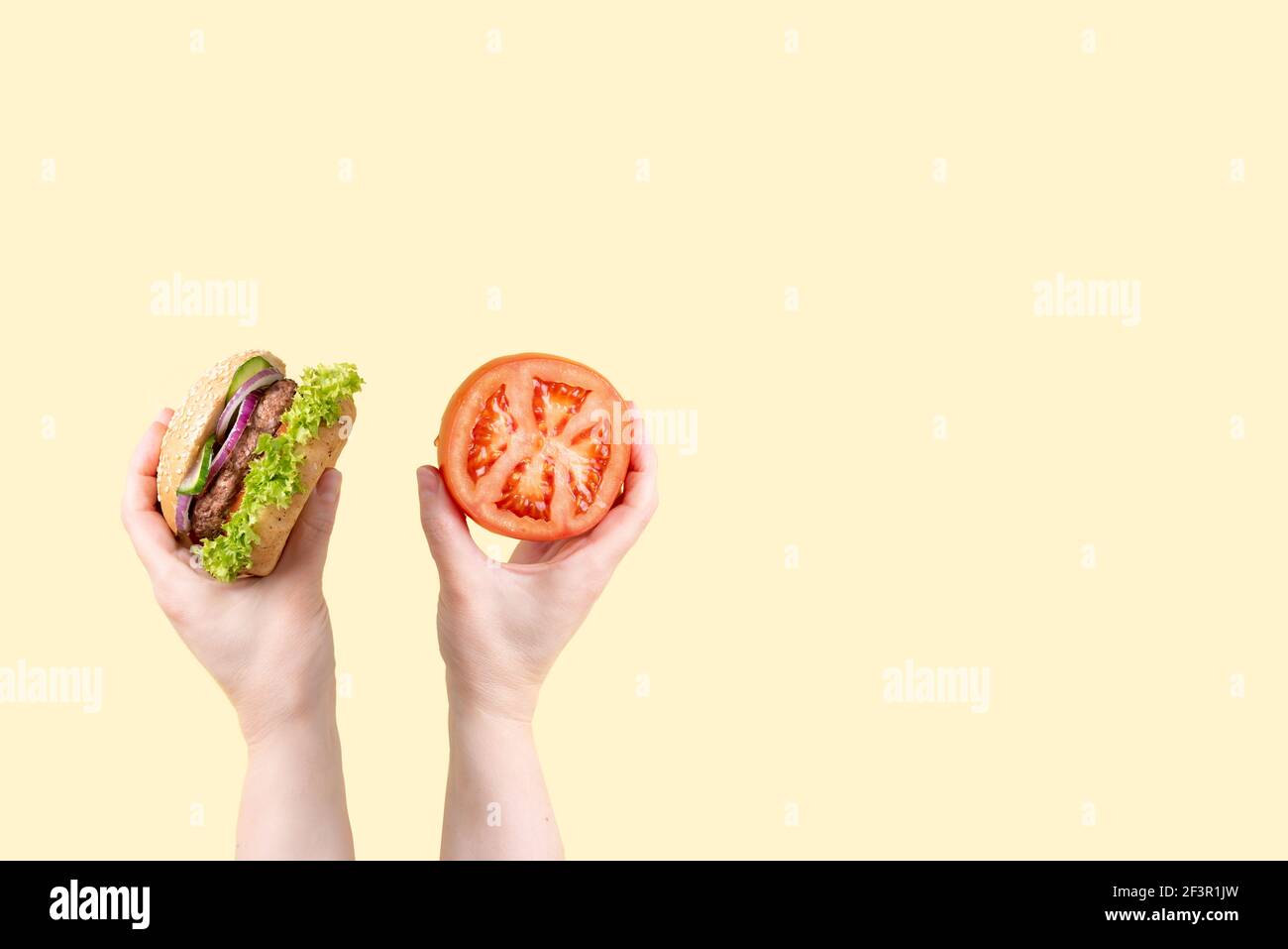 One female hand holding a burger, the other is holding a tomato on the yellow background, the choice of a way to eat concept. Stock Photo
