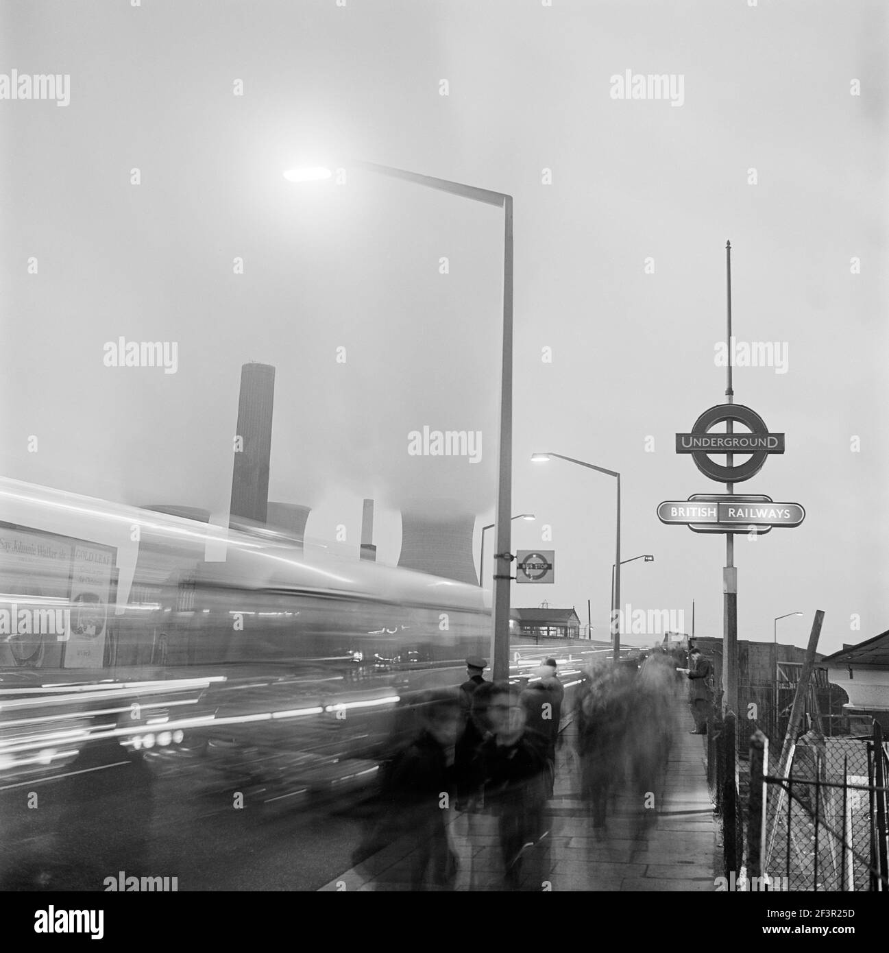 HARLESDEN, Greater London. A street scene with traffic and pedestrians passing a bus stop by a London Underground station sign in Harlesden at night, Stock Photo
