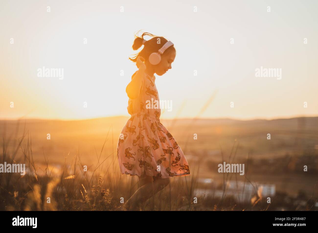 Side angle portrait of child with pink headphones and dress listening to music and dancing on a field at sunset Stock Photo