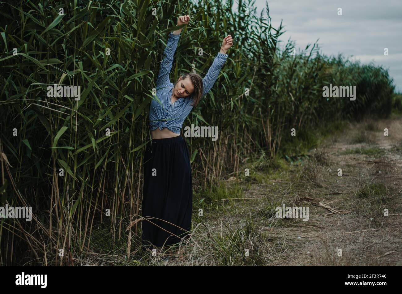 Full body artistic portrait of a woman with arms outstretched near red reeds on a path, moody vibe Stock Photo