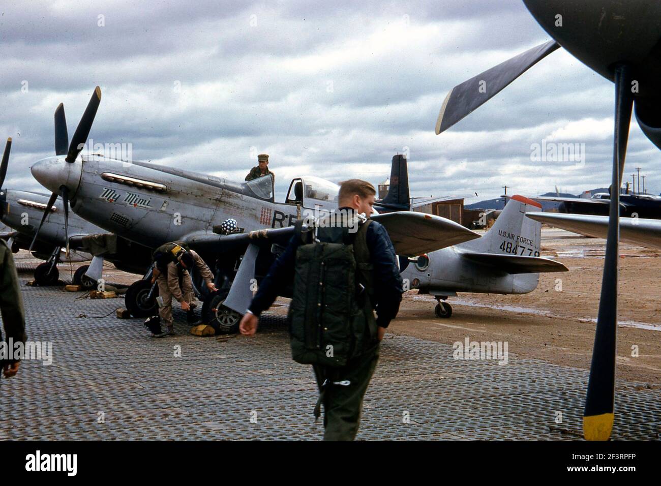 One-quarter left side view of a North American RF-51D 'My Mimi' (s/n 44-84778; c/n 44634) parked in the flight line somewhere in Korea. The pilot is shown walking towards the aircraft, and his helmet sits on the left wing. A crewman stands on the right wing beside the cockpit, and another bends down beside the left wheel. The propeller of another RF-51D Mustang is visible on the right side of the image, and two more appear on the left side. In the background is a Douglas B-26B Invader Stock Photo