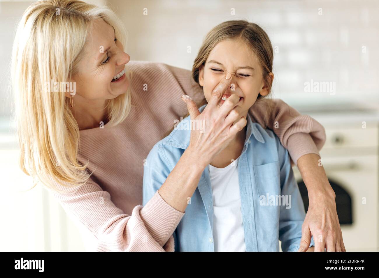 Beautiful caucasian middle aged blonde woman and preschooler girl in the kitchen. Grandma hugging granddaughter, touching her nose finger with flour, they have fun together at home Stock Photo