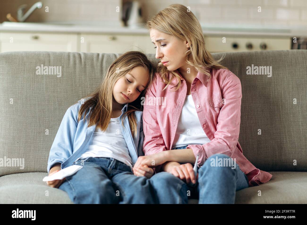 The woman and little girl are sitting at home on the couch. A young blonde mom in sits next to her sick and tired daughter, her head bowed on the shoulder of a worried mother Stock Photo