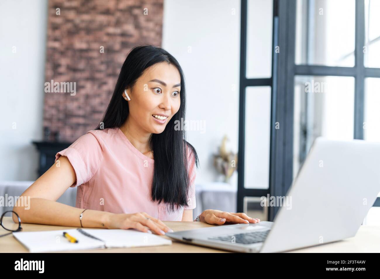Young Asian mixed-race woman with earphones sitting at the office desk, browsing internet and reading news online, looking at the laptop screen, cannot believe her eyes, feeling surprised shocked Stock Photo