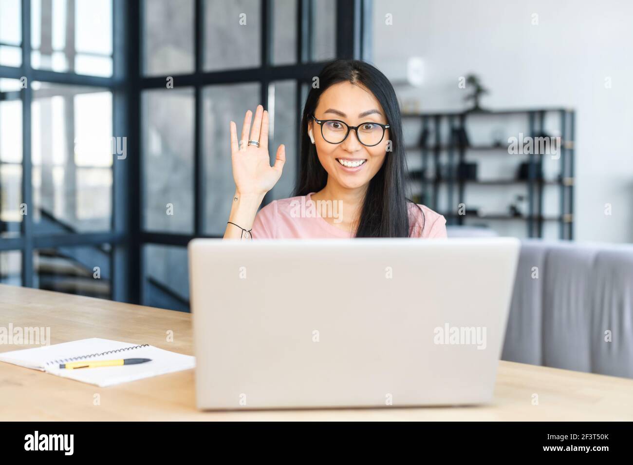 Intelligent Asian female manager in glasses sitting at desk in modern office, waving at the laptop, wearing earphones, mixed-race woman is talking to customer online, discussing project with client Stock Photo