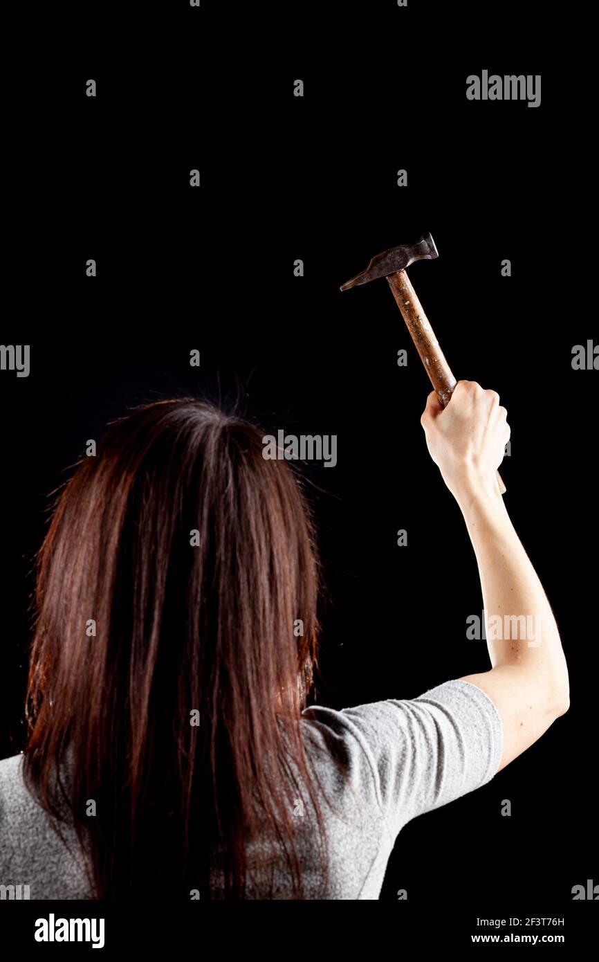 A rim light dark background image of a young caucasian woman with long brown hair. She is raising her right hand holding a hammer. A protest, activism Stock Photo