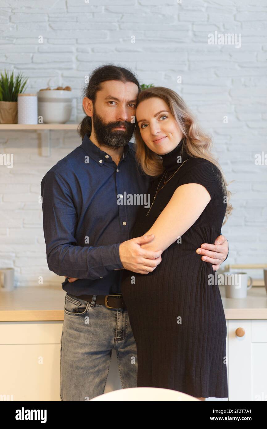 Man embracing pregnant partner in kitchen. Adult family pregnancy concept. Future parents in home outfit embrace standing in the kitchen, looking at e Stock Photo