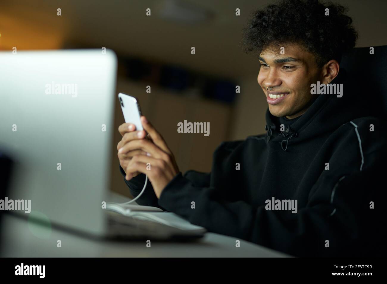 Distraction. Portrait of happy male student smiling and using smartphone while studying, working on laptop late at night Stock Photo