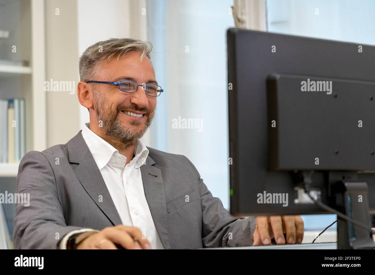 Successful Mature Businessman Sitting at Office Desk. Smiling Elegant Man Looking at Computer Monitor in His Office. Stock Photo