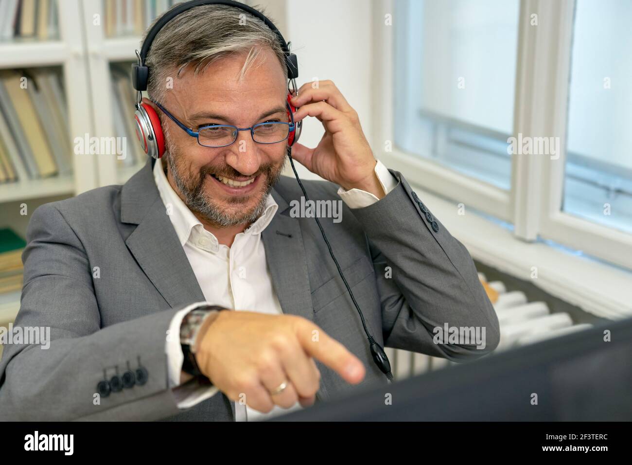 Smiling Mature Businessman With Headphones Sitting at Office Desk Pointing at Computer Monitor. Excited Businessman Gesturing with Hand in His Office. Stock Photo
