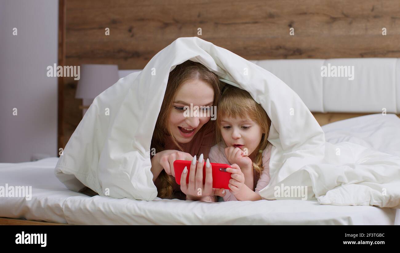 Mother lying on bed under duvet blanket with daughter kid girl holding