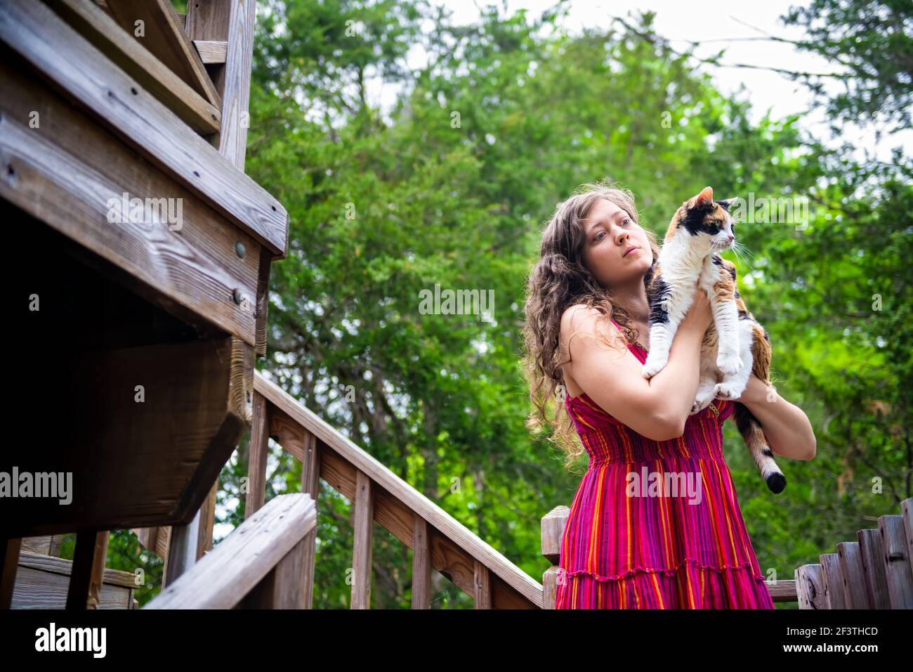 Happy smiling young woman bonding holding in hands calico cat pet companion, friends showing affection outside outdoors in home garden on wooden deck Stock Photo