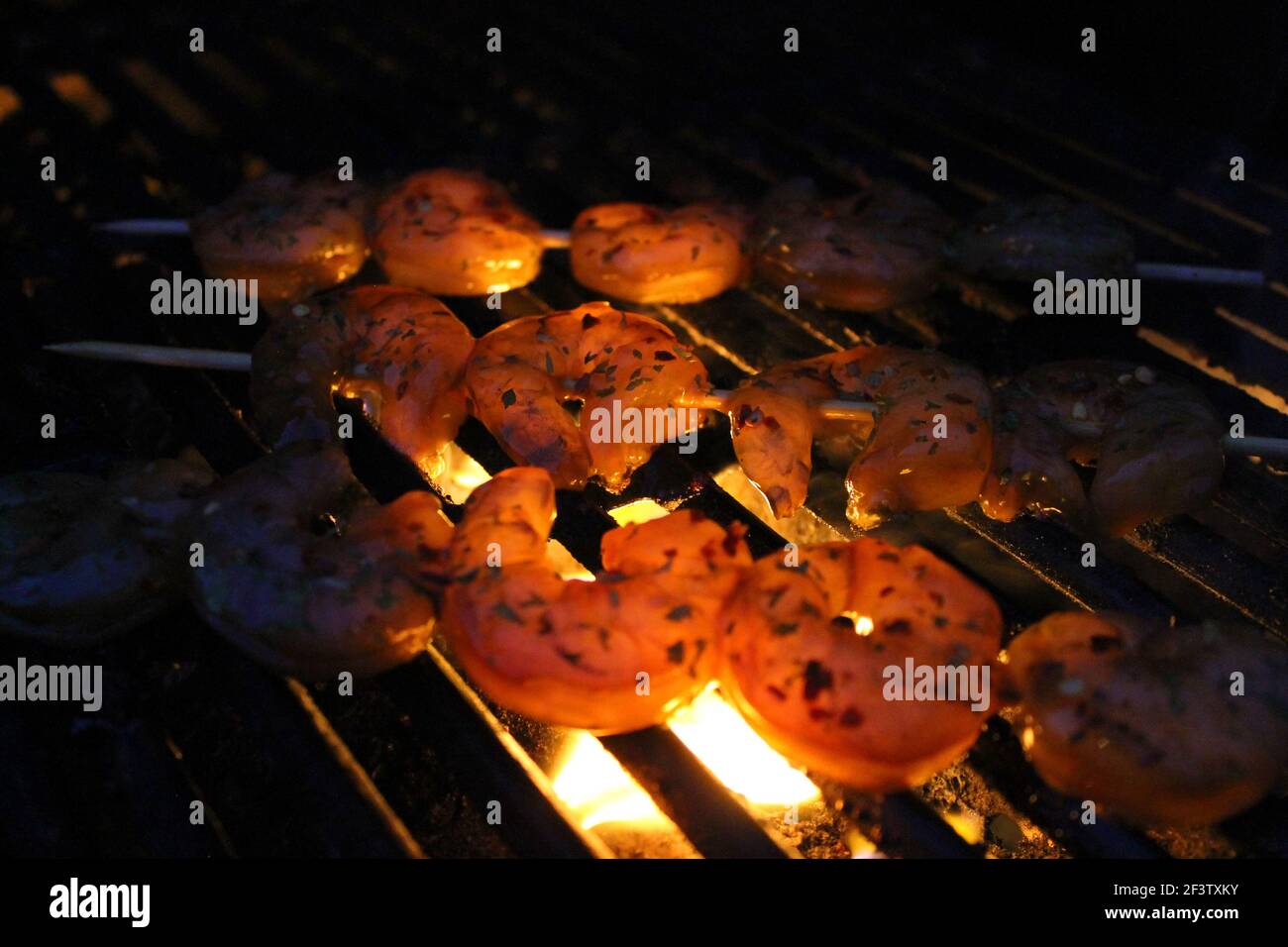 Close-up of shrimp on a barbecue grill. Lighting is from flame below shrimp, bright in center, darkening around the edges. Shrimp are on Skewers. Stock Photo