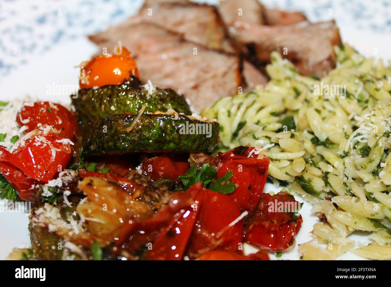 Close-up of a plate of fried cherry tomatoes and fried zucchini with basmati rice, and sliced baked pork, out of focus in background. Stock Photo