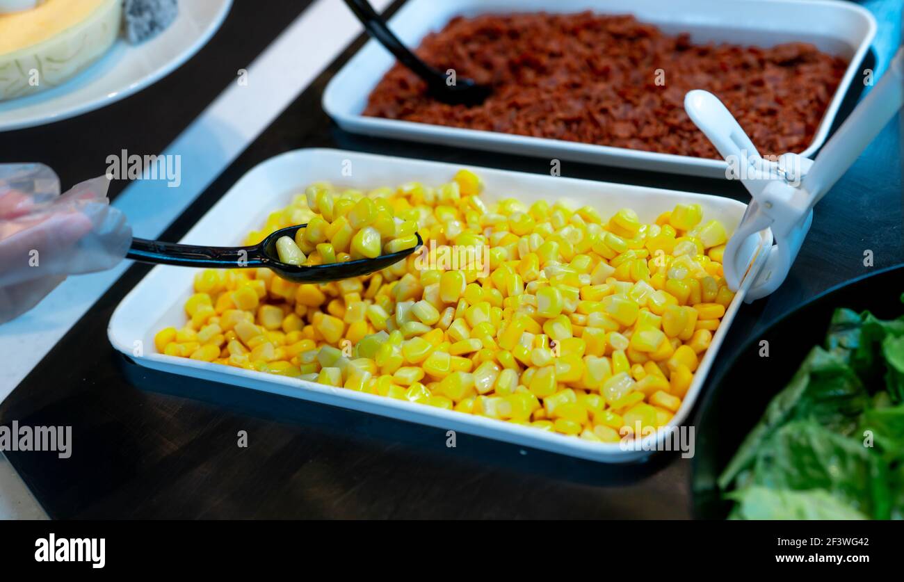 Selective focus on sweet corn in white bowl at salad bar buffet of restaurant. Hand wear plastic gloves holding black spoon scoop yellow sweet corn. Stock Photo