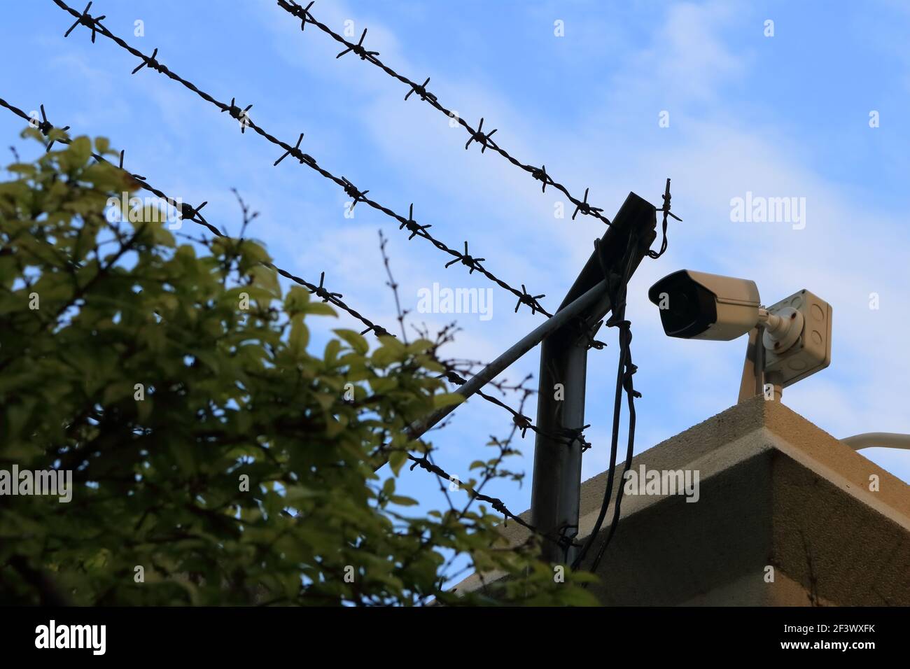 Closeup of security camera installed on rooftop next to barbed wire against blue sky background Stock Photo