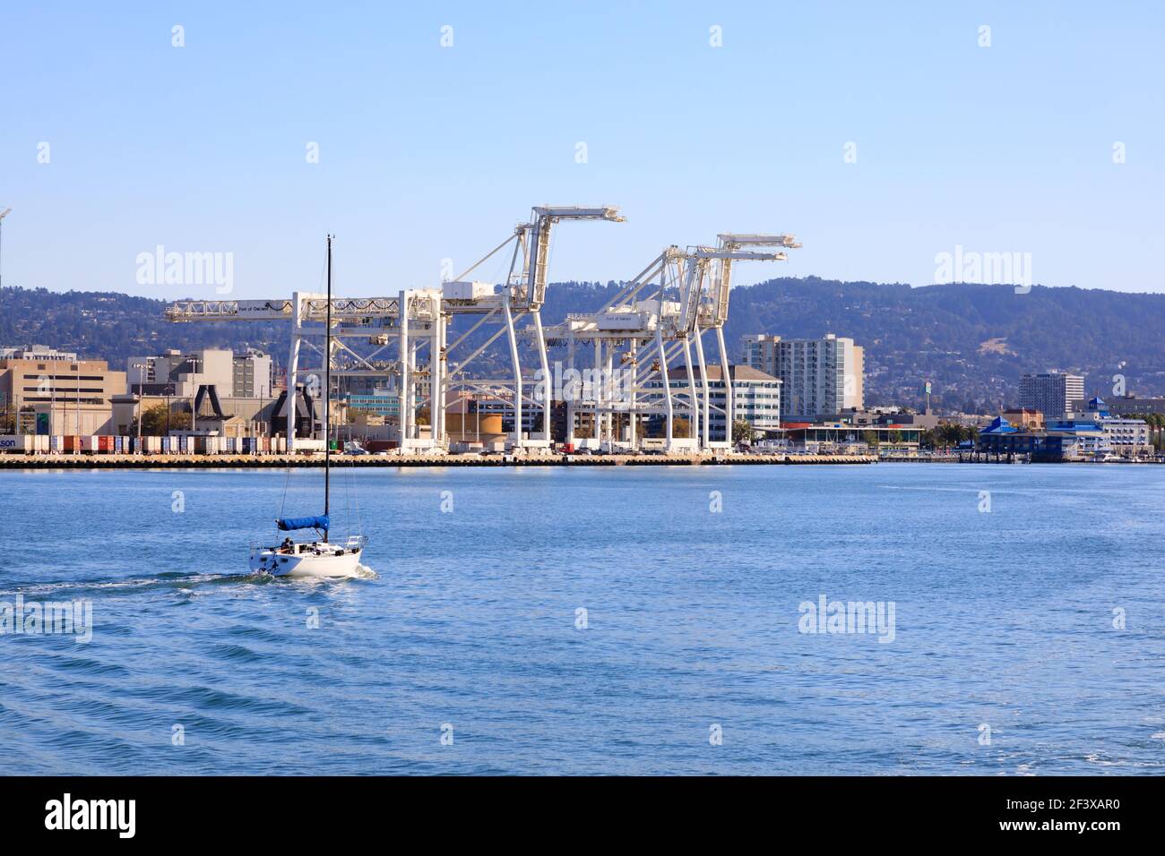 A lone yacht motors towards Jack London Square, past the cranes of the Port of Oakland, San Francisco, United States of America. Stock Photo