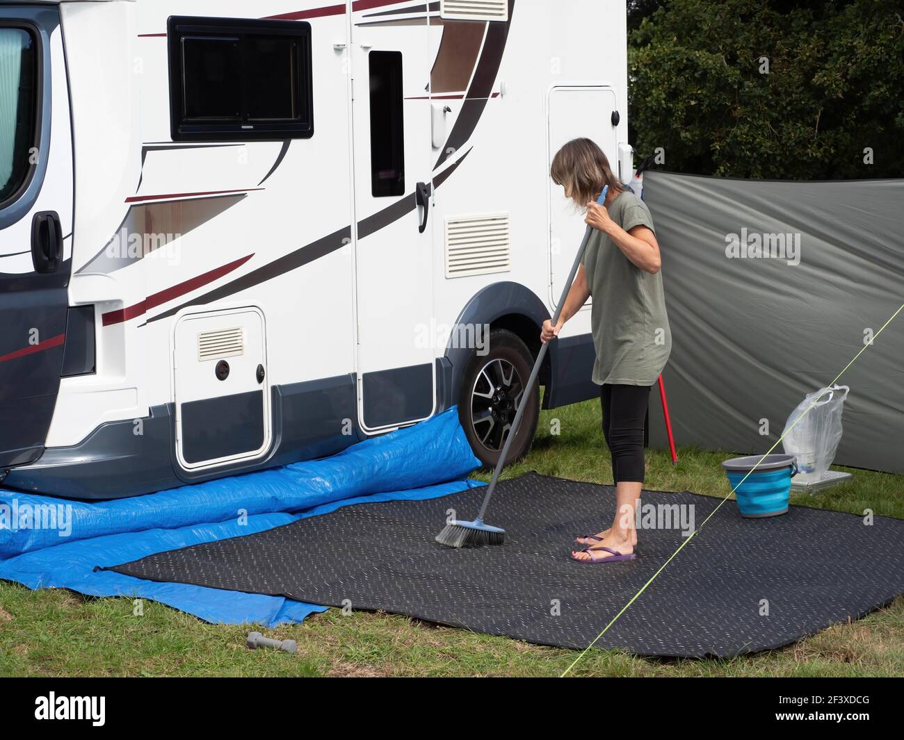 A lady motorhome owner sweeps her outdoor mat with a broom outside her recreational vehicle. A bucket and wind break are visible Stock Photo