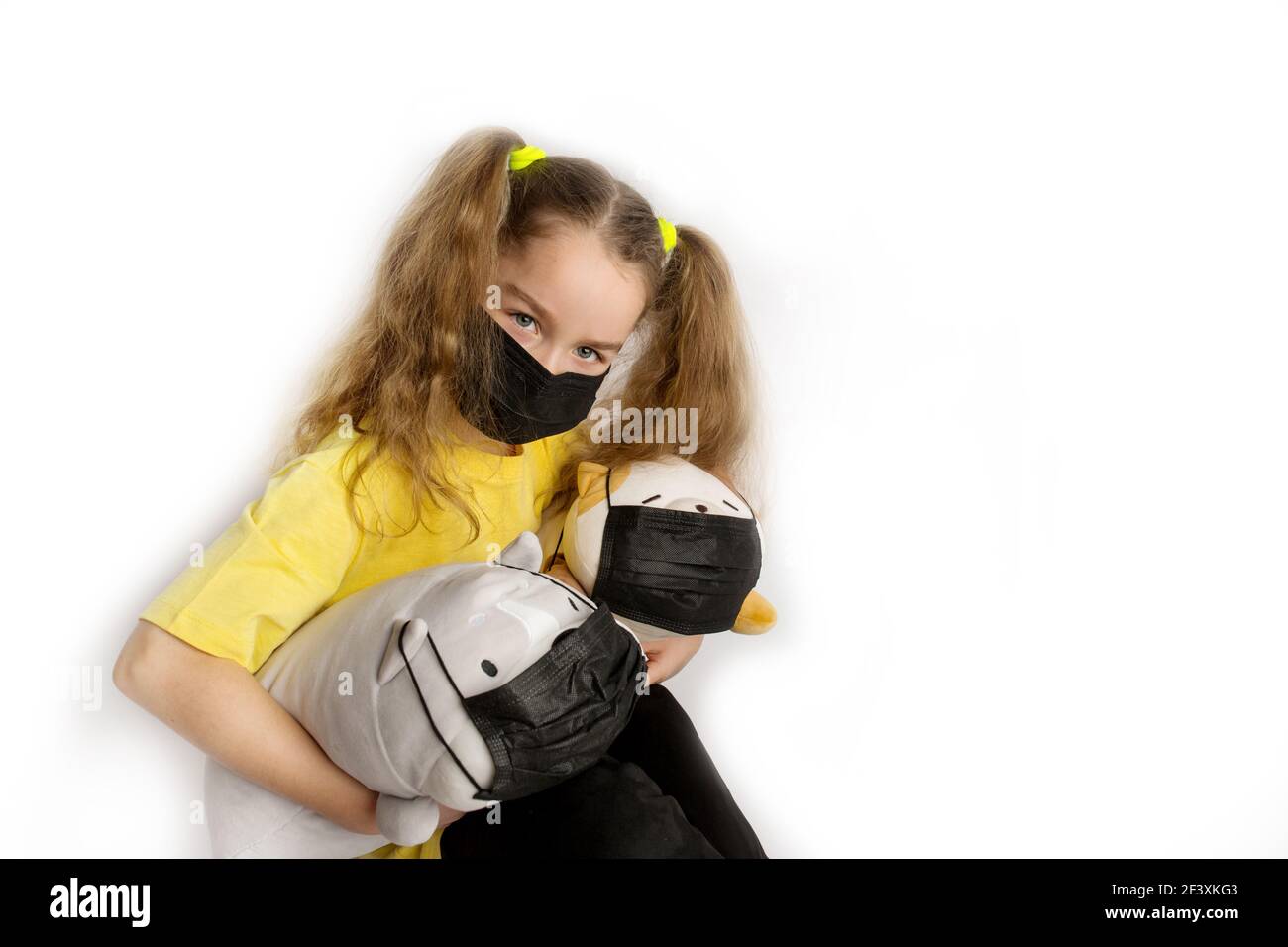Girl in a yellow t-shirt and black mask covid19 on a white background with toys in a black covid mask Stock Photo