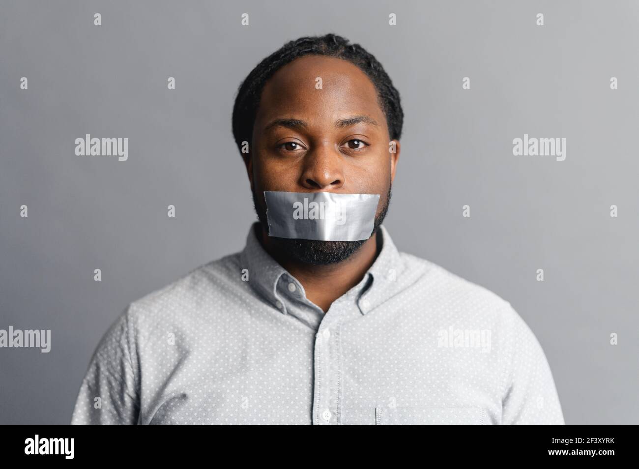 Serious African-American man with with a sealed mouth with tape looks at the camera sadness, speechless, speechless, racism concept, isolated on grey Stock Photo