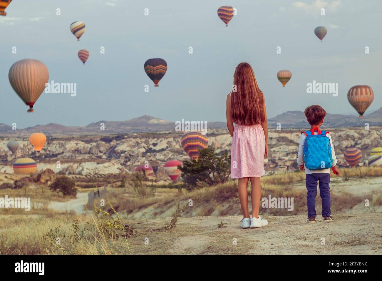 two kids girl in a pink dress and a boy with backpack look on multicolored balloons in the sky above the sandy mountains Stock Photo