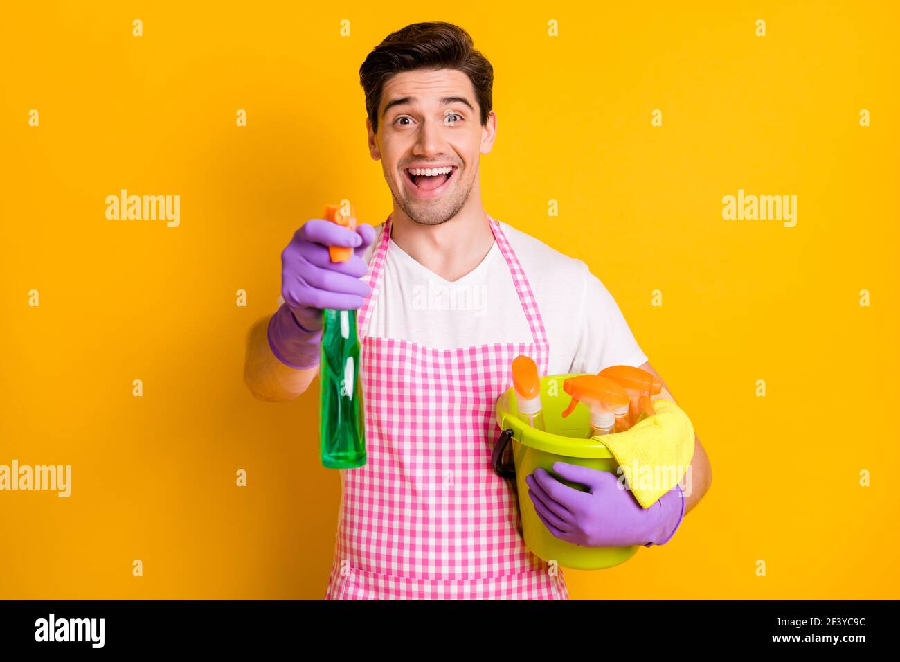 Photo of young happy excited smiling man in gloves hold cleaning splay and bucket with detergent isolated on yellow color background Stock Photo